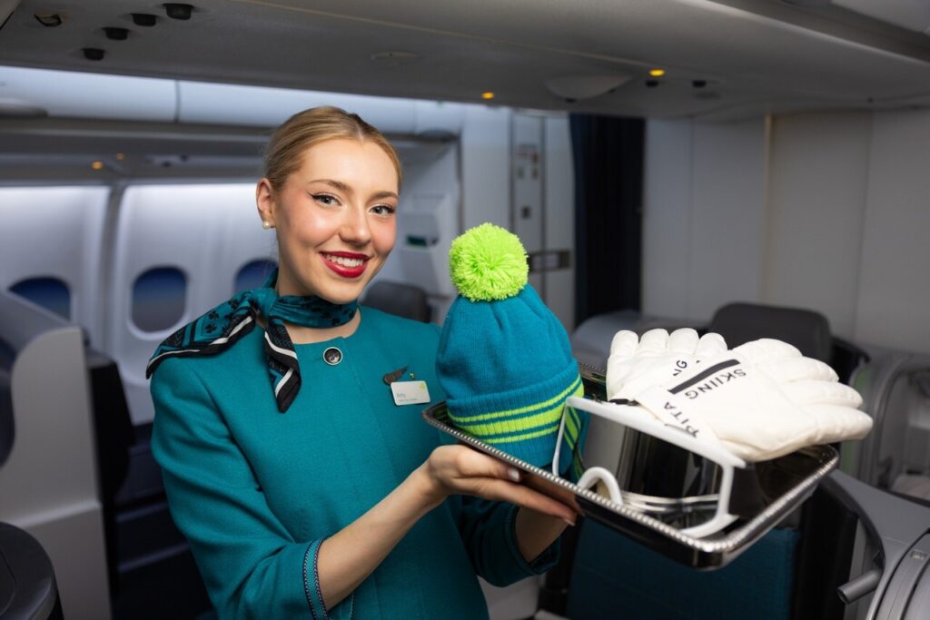 Female Aer Lingus cabin crew member in an airplane. She's holding a tray with a hat, ski gloves and ski goggles.